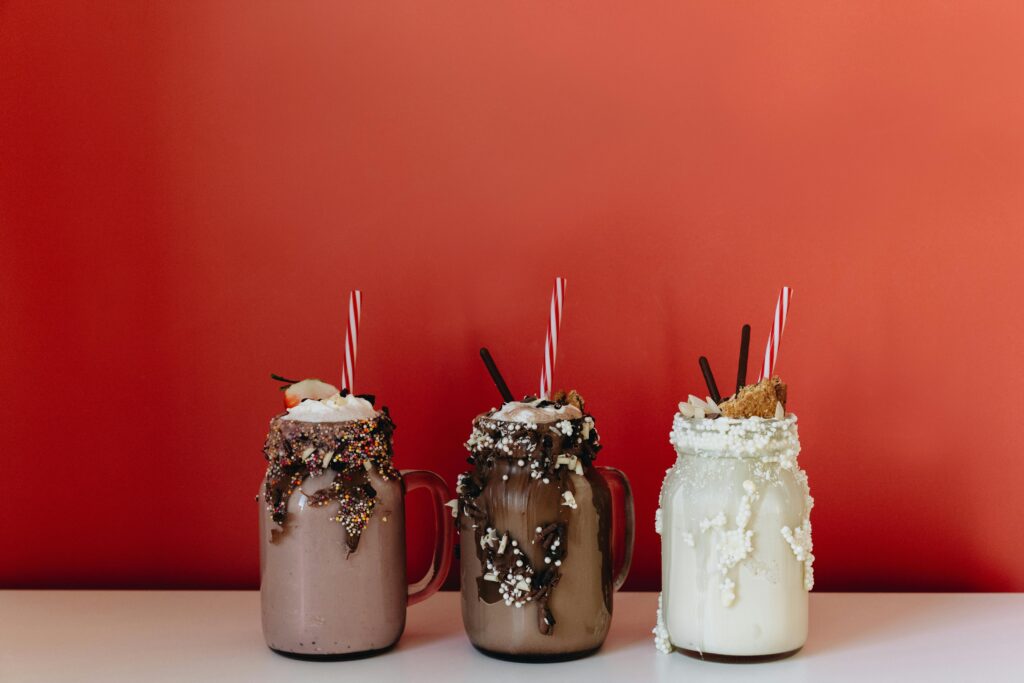 Three indulgent milkshakes in glass jars with sprinkles and chocolate against a bold red backdrop.