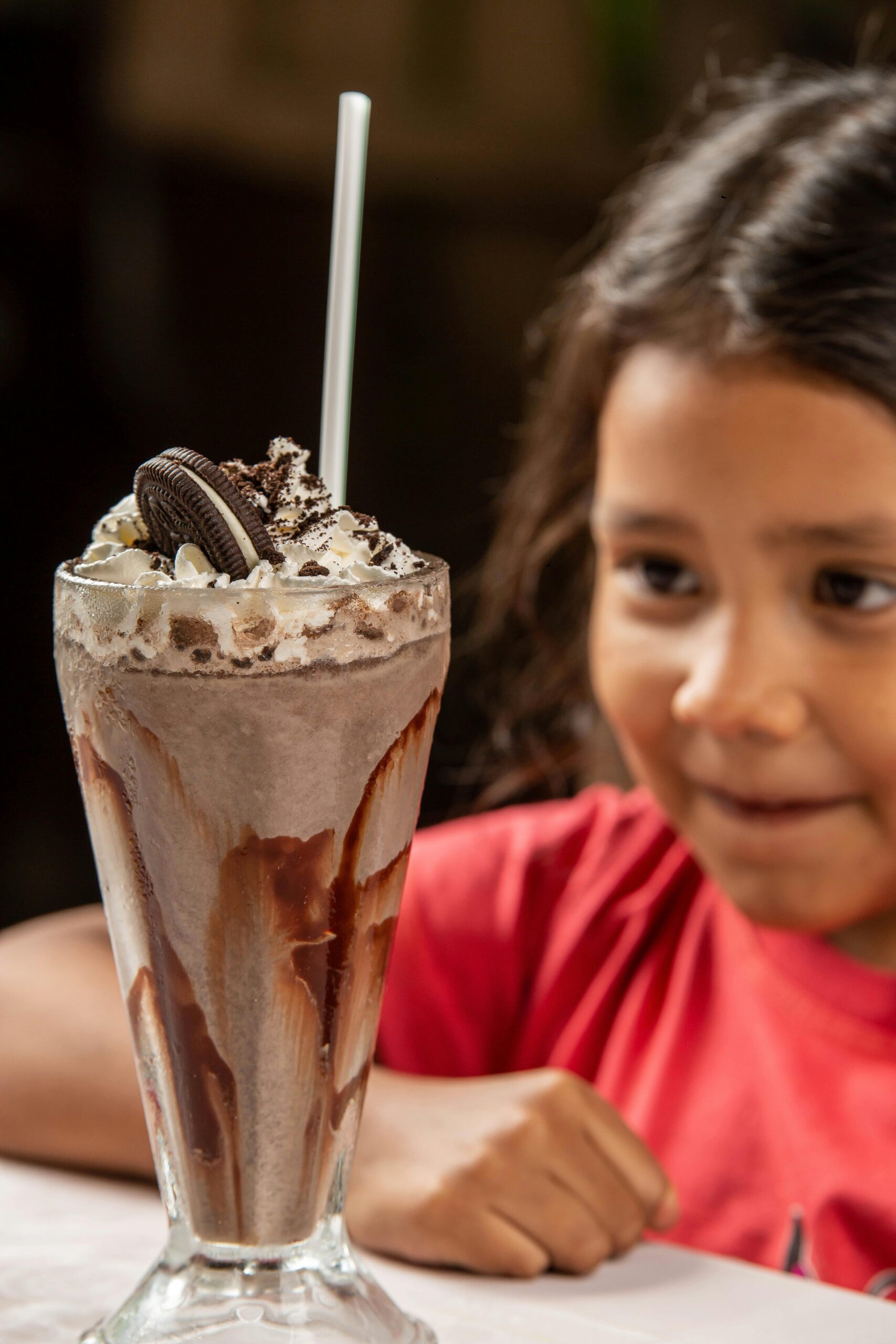 A child excitedly looks at a chocolate milkshake with whipped cream in a glass cup.