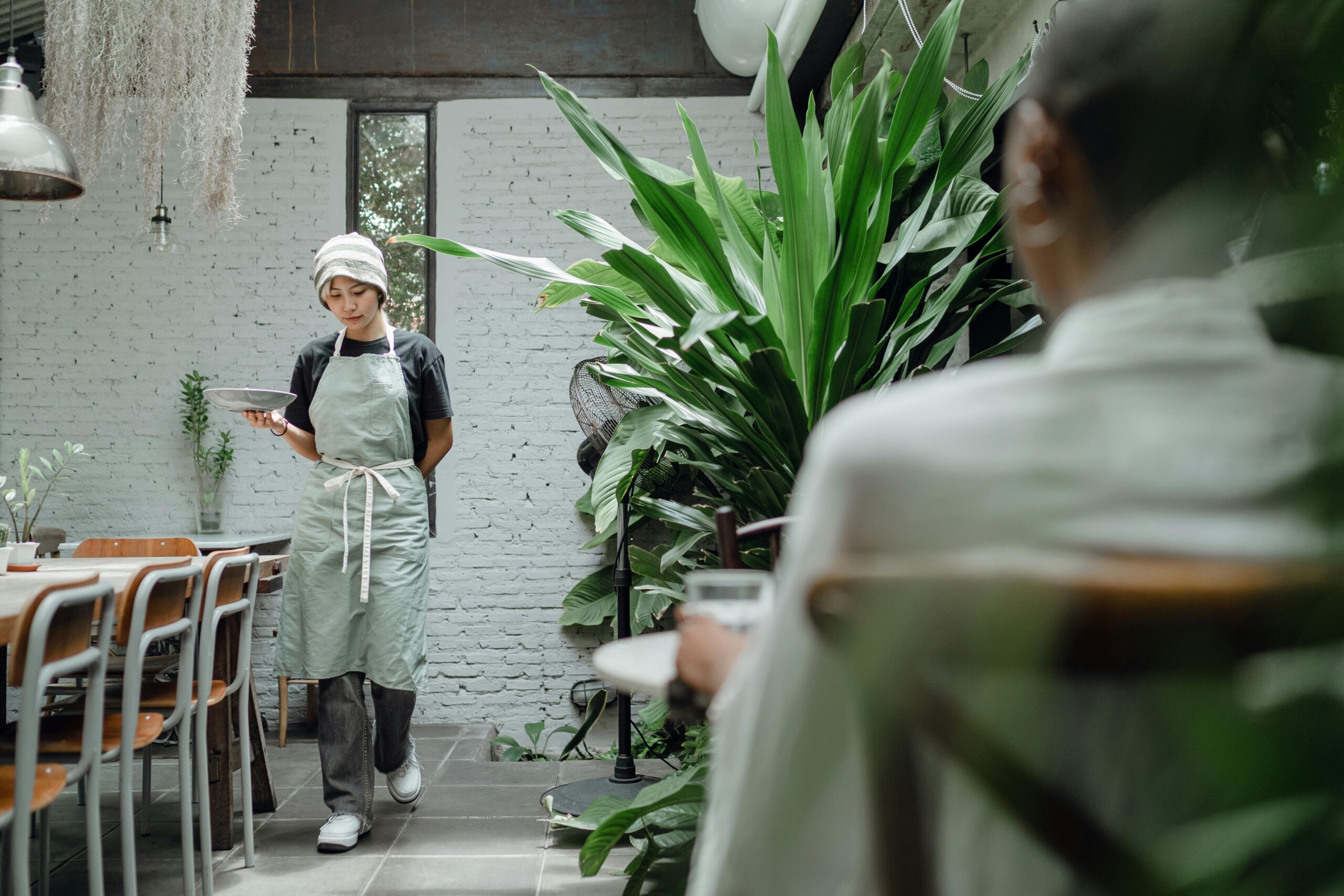 Woman in a modern restaurant setting carrying food, surrounded by lush greenery.