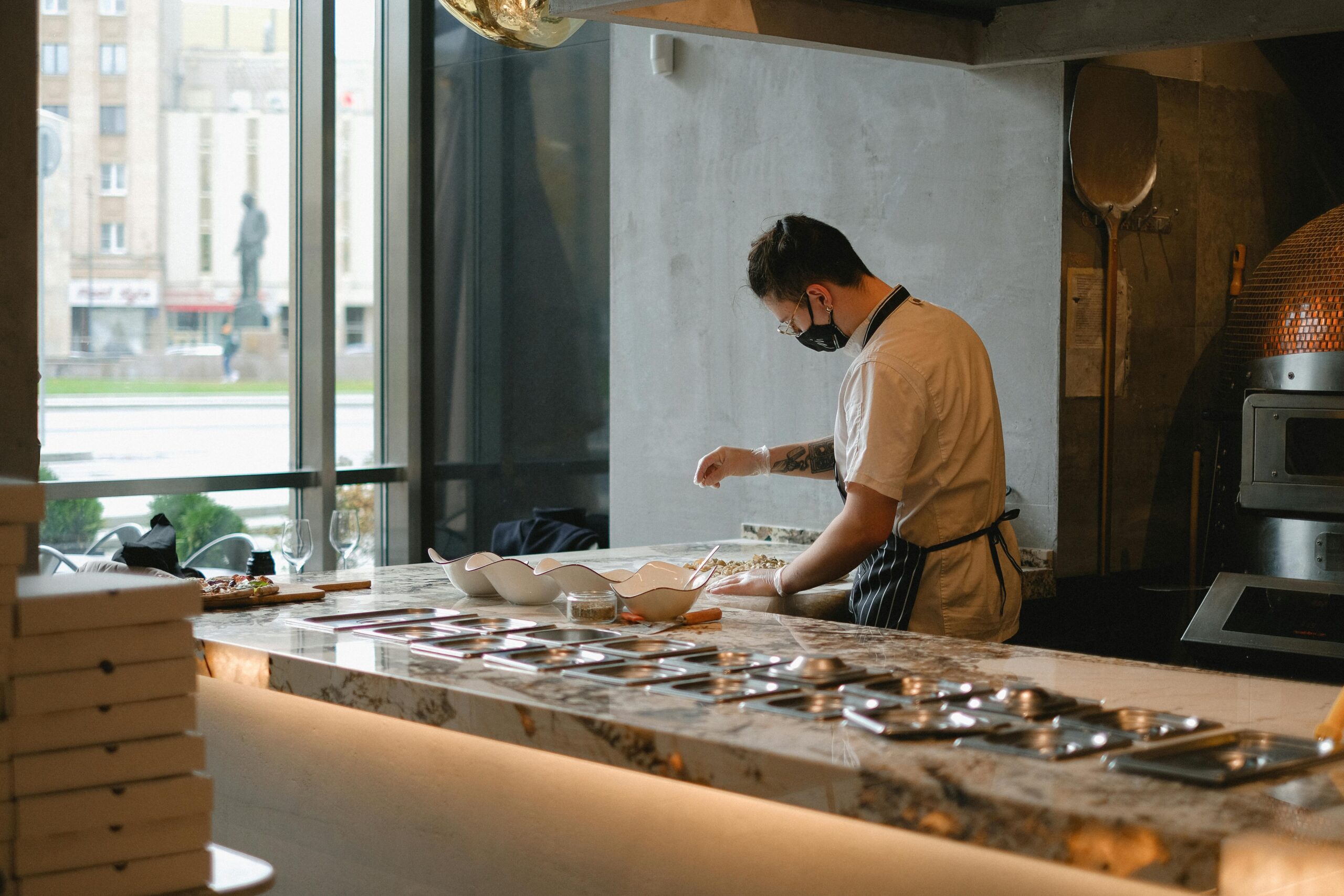 A chef wearing a mask prepares pizza dough in a stylish pizzeria with a wood-fired oven.